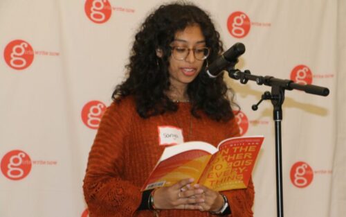 Sanya, a girl with curly hair, reads from the 2023 anthology, On the Other Side of Everything, at a microphone; GWN step and repeat in the background