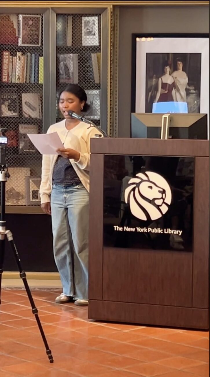 Madison Mailutha, a young woman with dark skin and black hair, reads from a piece of paper at a podium at NYPL